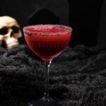 A blood beet red mocktail sits in a tall glass with black salt on the rim and a skull sitting in the background.