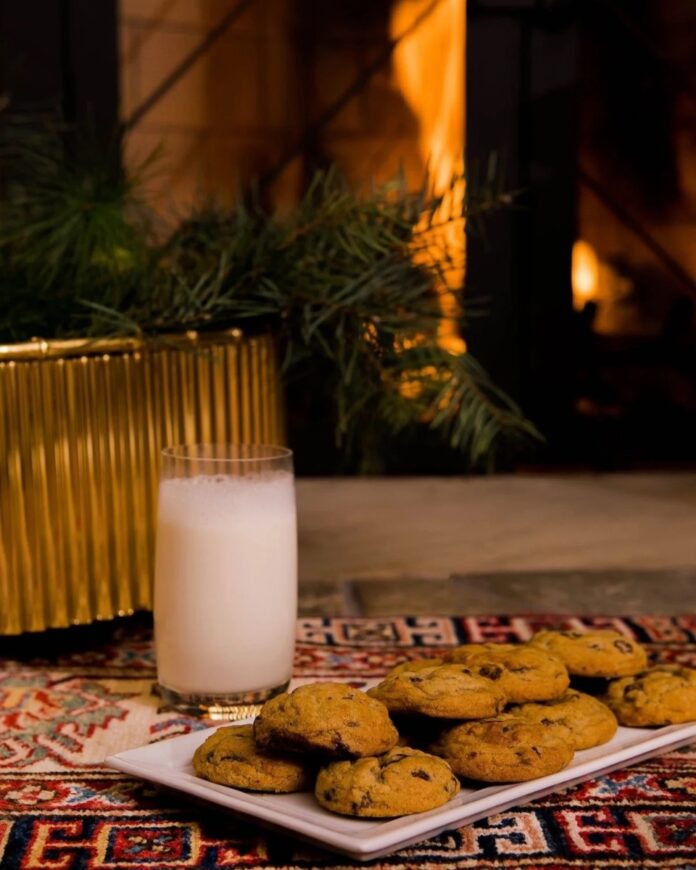 A plate of warm chocolate chip cookies and a glass of milk on a festive rug, with pine branches and a fireplace glowing in the background.