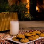 A plate of warm chocolate chip cookies and a glass of milk on a festive rug, with pine branches and a fireplace glowing in the background.