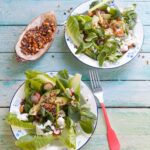 Two plates of green salad with romaine, avocado, goat cheese, and radishes, topped with a savory Cacao Dukkah, served on a rustic blue wooden table next to a small wooden boat of the spice blend.