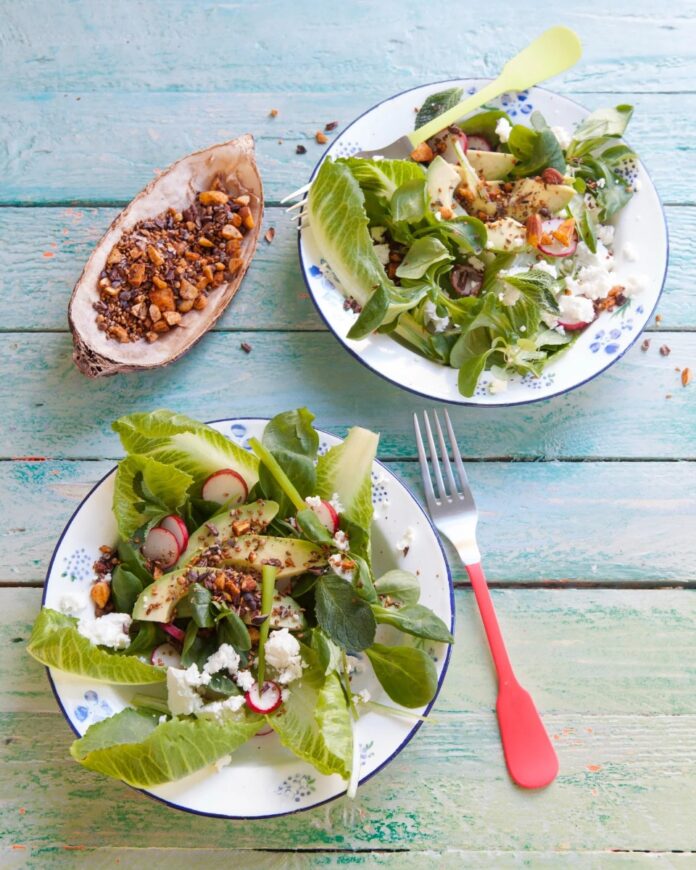 Two plates of green salad with romaine, avocado, goat cheese, and radishes, topped with a savory Cacao Dukkah, served on a rustic blue wooden table next to a small wooden boat of the spice blend.