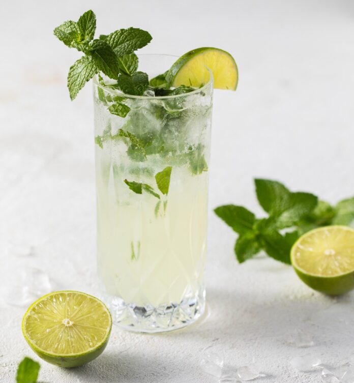 A tall glass with a Classic Mojito inside, garnished with mint and a life wedge, sits on a white background with half limes and sprigs of mint on the table as well.
