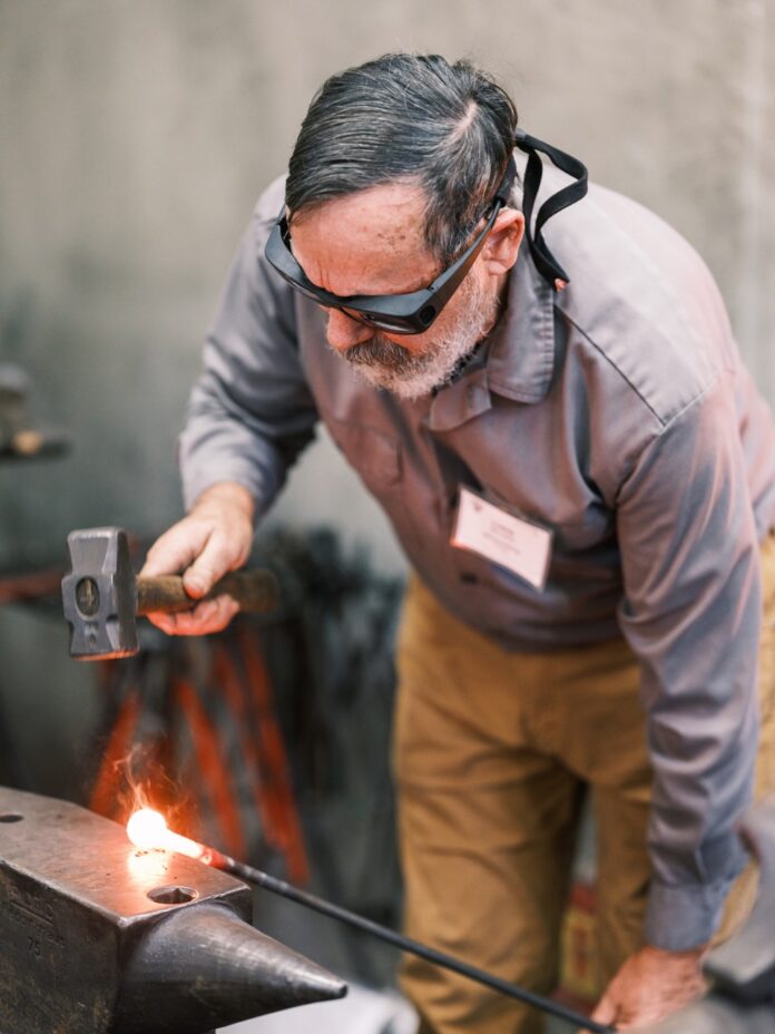 A strikes a hot piece of metal with a hammer as he wears craft safety goggles.
