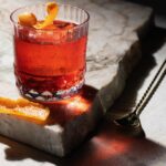 A craft negroni made with rum sits on a marble block with an orange peel garnish inside the short glass and a spoon beside the marble block.