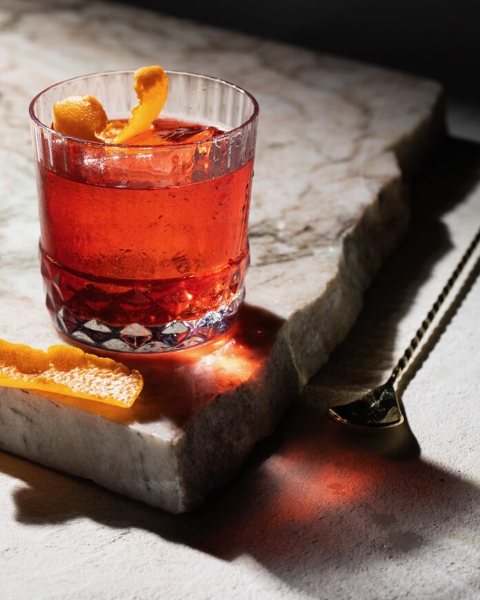 A craft negroni made with rum sits on a marble block with an orange peel garnish inside the short glass and a spoon beside the marble block.