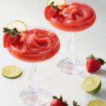 Two tall glasses hold strawberry daiquiris, frozen and topped with strawberries and lime slices as more strawberry and lime slices lay on the table below.