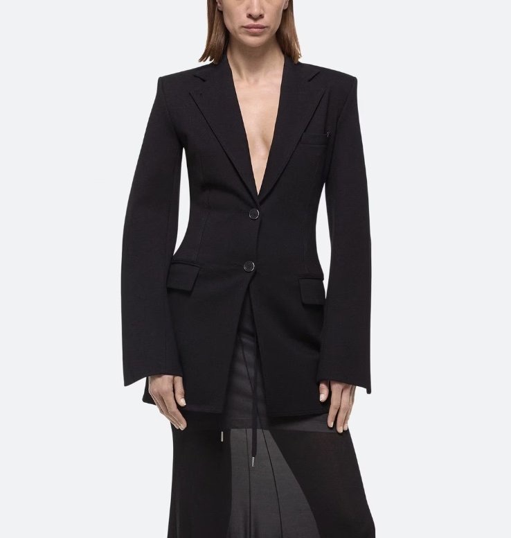 A woman in a black long blazer stands in front of a white background.