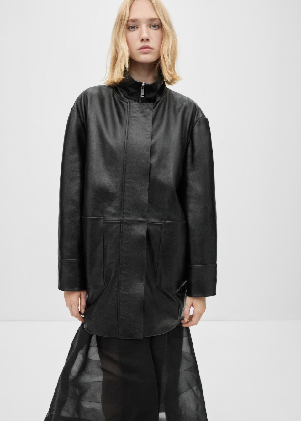 A woman stands in a long leather, fall trench coat and skirt in black.