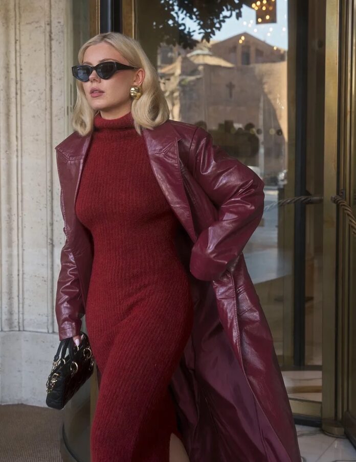 A woman in a red fall jumpsuit and red leather trench coat walks with a hand in her pocket.