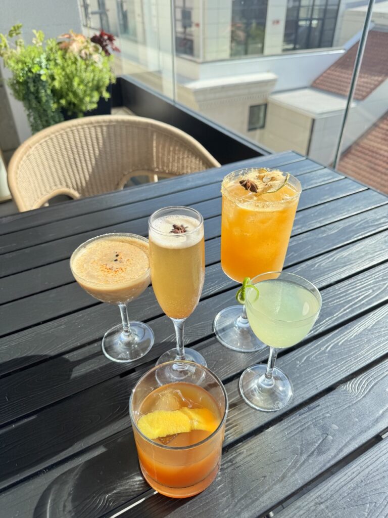 5 fall cocktails sit on a table outside in the sun in various shades of orange.