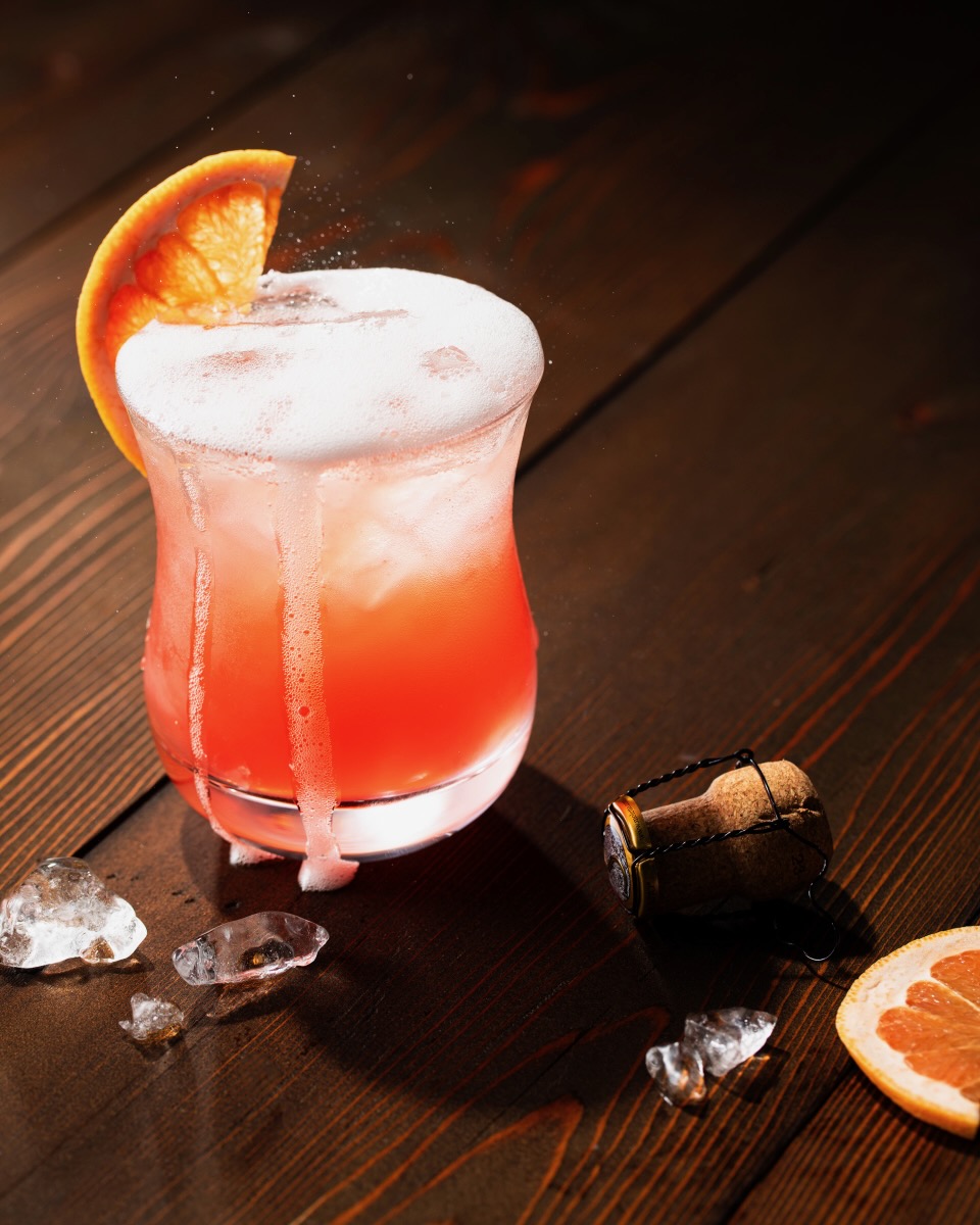 A ruby red Italian Greyhound cocktail sits in a glass, bubbling over from a splash of prosecco and topped with a grapefruit slice. The cork from the bottle, ice, and another grapefruit slice sit on the dark wood table nearby.