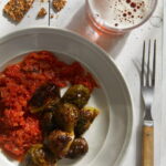 A white dish on a white surface with red kimchi and roasted Brussels sprouts with a serving fork, a cup of beer, and some broken seeded crackers above the bowl.