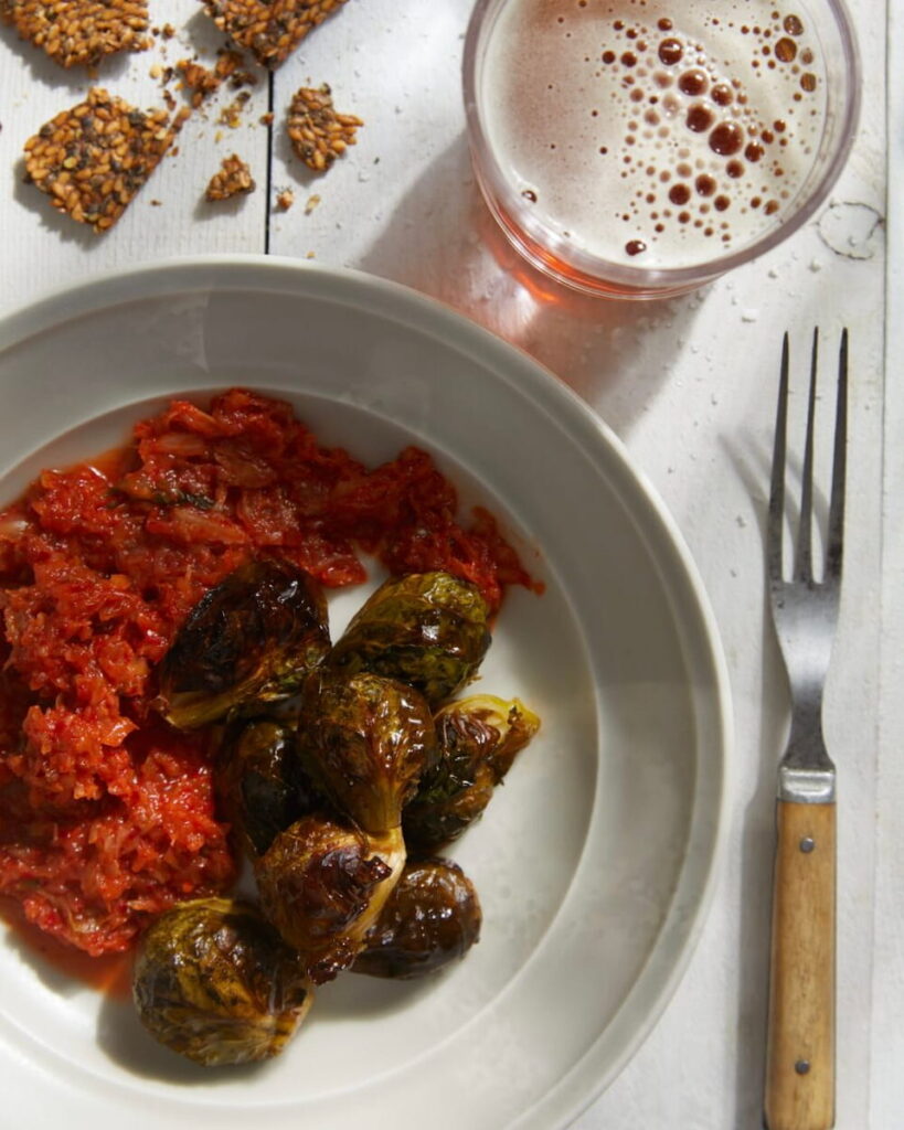 A white dish on a white surface with red kimchi and roasted Brussels sprouts with a serving fork, a cup of beer, and some broken seeded crackers above the bowl.