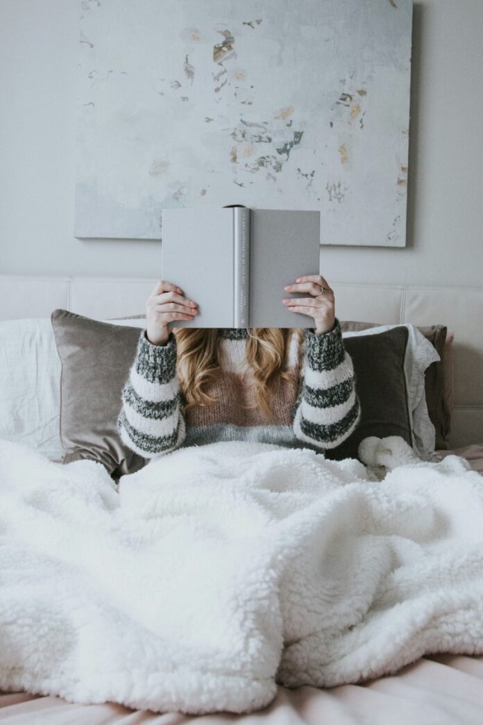 October Book Releases to Add to Your TBR A woman in a sweater sits under a white blanket with a beige book in front of her face.