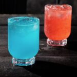 A red cocktail sits slightly behind a blue cocktail both in the same glasses.