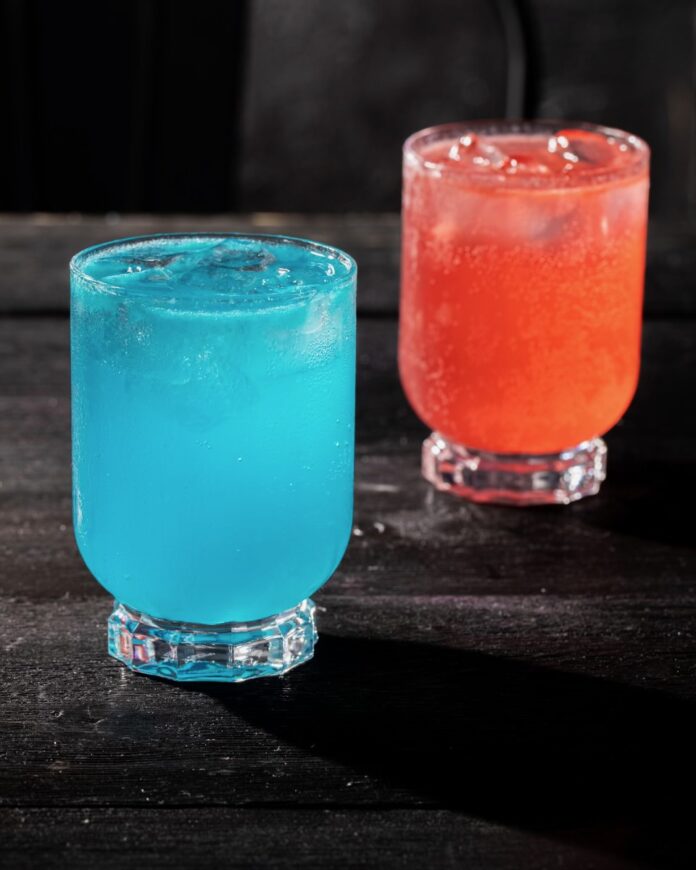 A red cocktail sits slightly behind a blue cocktail both in the same glasses.