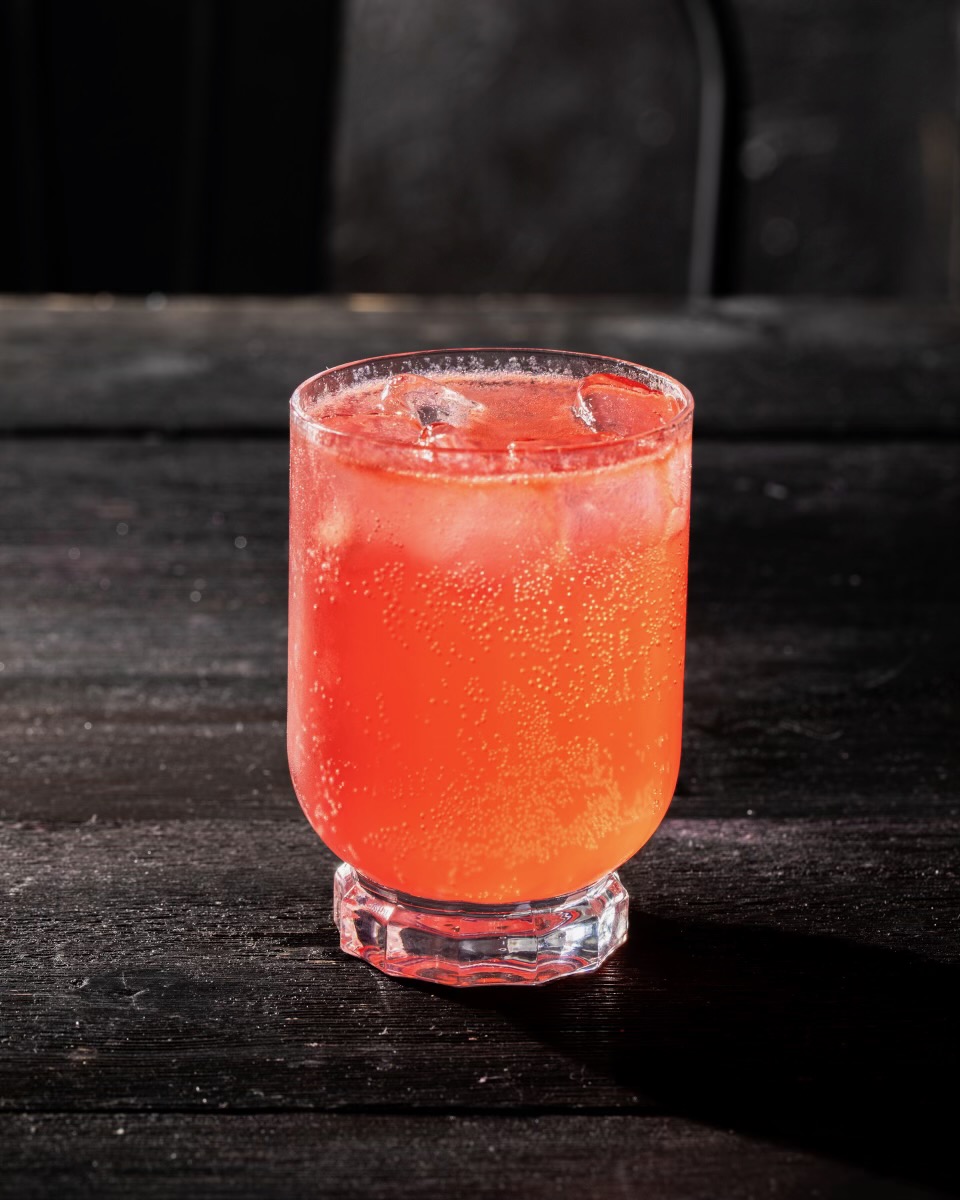 A red cocktail in a short glass sits against a black background. 