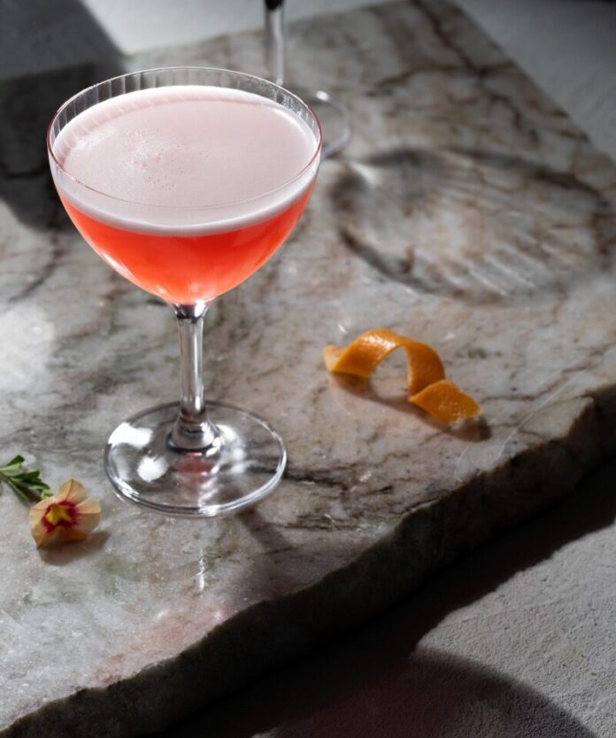 In a tall cocktail glass sits a ruby red cocktail with the color of ruby red grapefruit juice and an egg white foam on top. It sits on a marble block with a grapefruit twist and elderflower nearby.