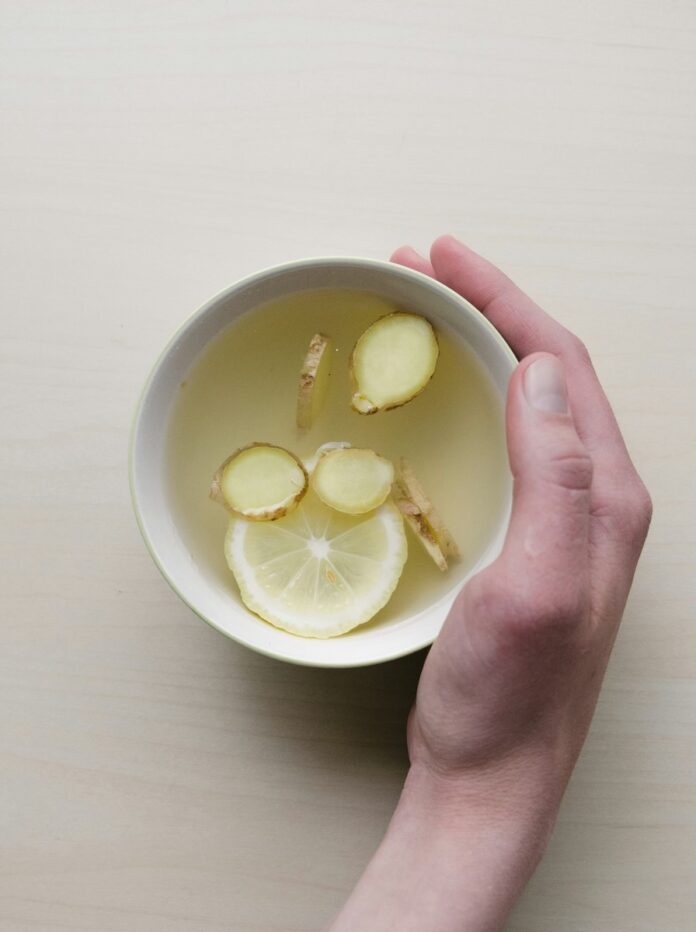 A person places their hand on a cup of ginger tea with slices of ginger and lemon sitting inside.