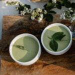 Two green bowls of soup laid on a rustic cutting board and arranged with flowers.