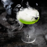 A green cocktail on a black background, styled for Halloween with spiders and smoke surrounding it
