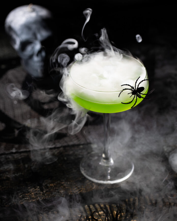 A green cocktail on a black background, styled for Halloween with spiders and smoke surrounding it
