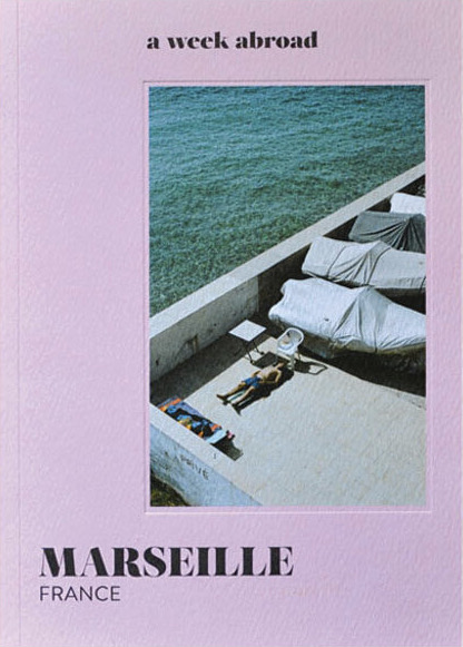 The cover of A Week Abroad: Marseille, pink with a black and white image on it of a man laying by the ocean in the South of France