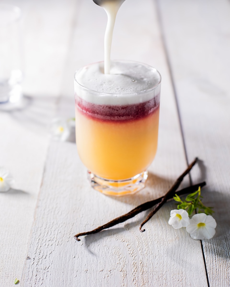 A short cocktail glass sits in the center of a white wood table with a New York Sour inside in the colors orange, red, and a white foam.