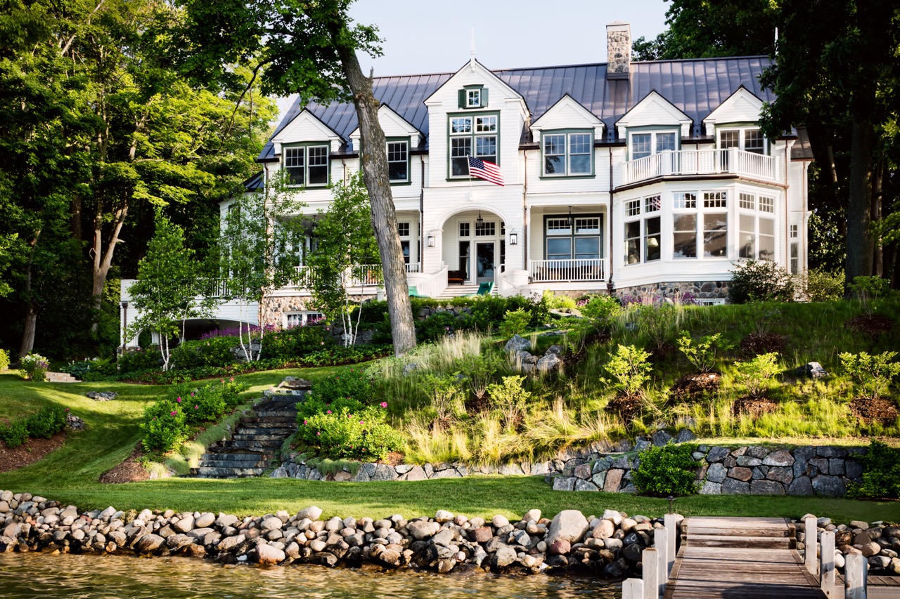 The outside of a huge white bouse in front of a lake with greenery all around. 