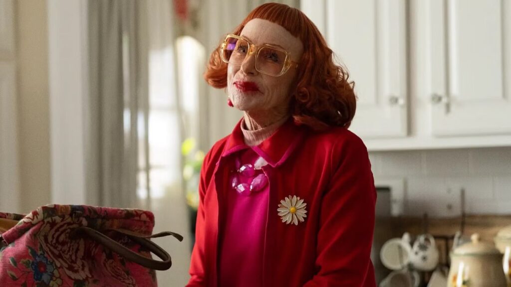 A woman with red hair, glasses, and pink clothing stands in a kitchen with bright red lipstick.