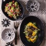 Two black dishes, one with a salad and one with a Bobotie over peas, sit on a tablecloth with flower decor around them.