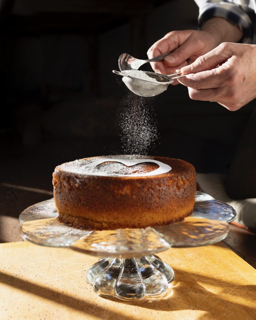 A Campari Olive Oil Cake sits on a glass platter as a person sprinkles powdered sugar in a heart shape over top.