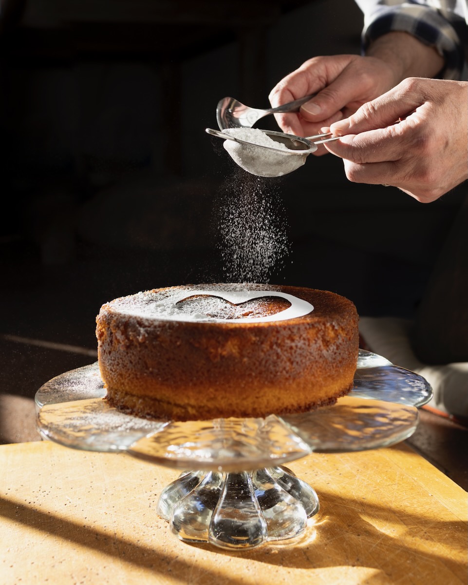 A Campari Olive Oil Cake sits on a glass platter as a person sprinkles powdered sugar in a heart shape over top.