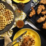 Southern fried chicken and waffles on bright yellow plates, drizzled with hot honey butter sauce, alongside a wooden cutting board with plain waffles.