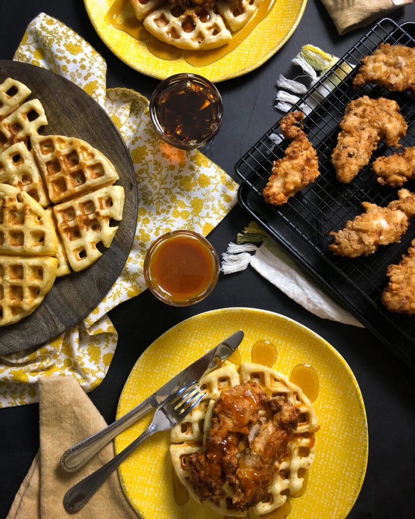 Southern fried chicken and waffles on bright yellow plates, drizzled with hot honey butter sauce, alongside a wooden cutting board with plain waffles.