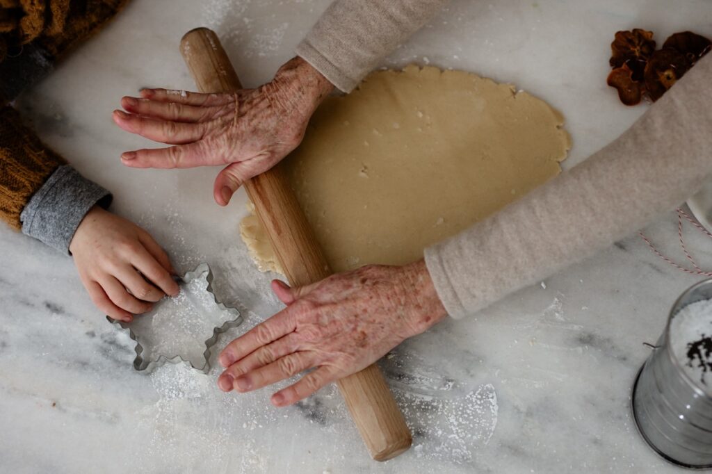 An older pair of hands rolls out cookie dough while young hands play with a snowflake cookie cutter.