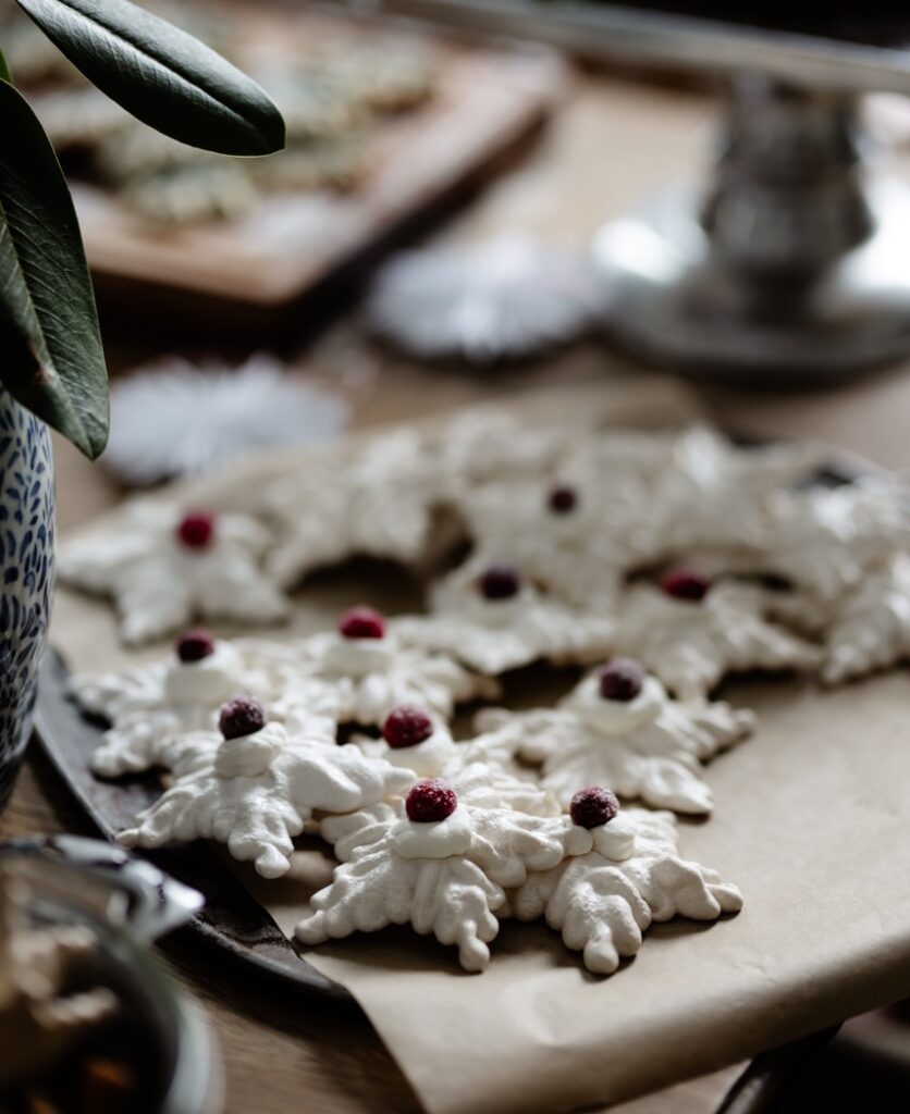 A plate full of French meringue cookies in snowflake shapes and topped with cherries amongst a table full of cookies.