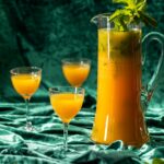 A large glass pitcher filled with a vibrant orange Piña Paradise Mojito cocktail garnished with mint, beside two glasses ready to serve.