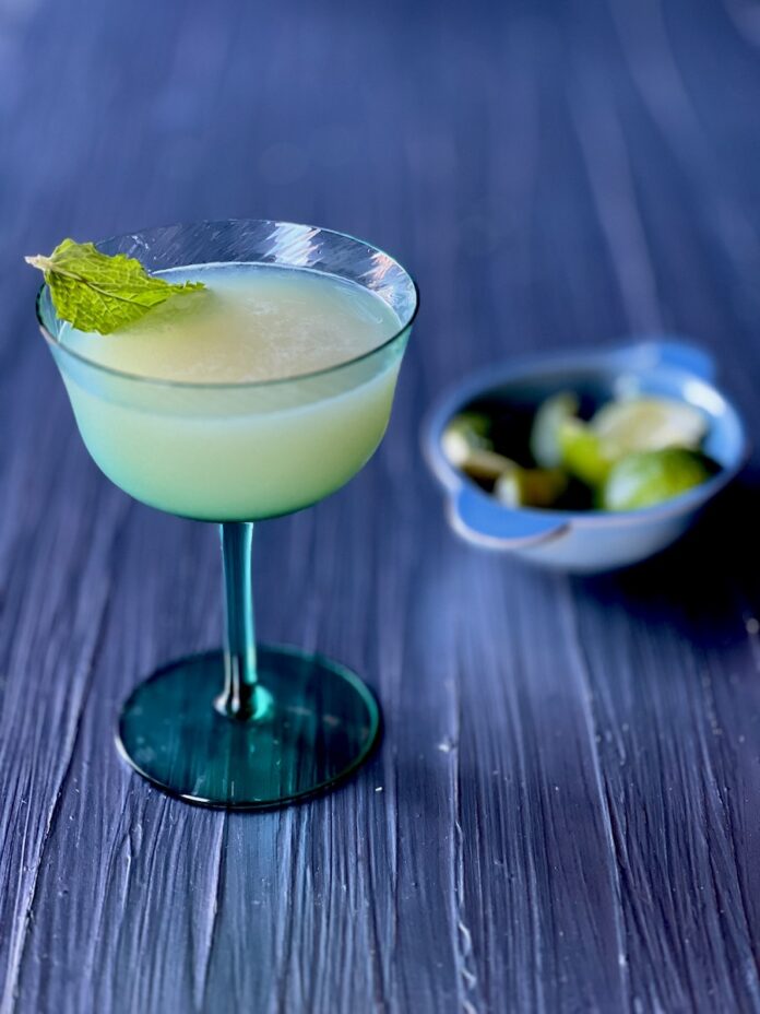 A coupe glass holds a gin and lime juice combination with a mint leaf garnish as a bowl of lime wedges sits to the right of the glass.