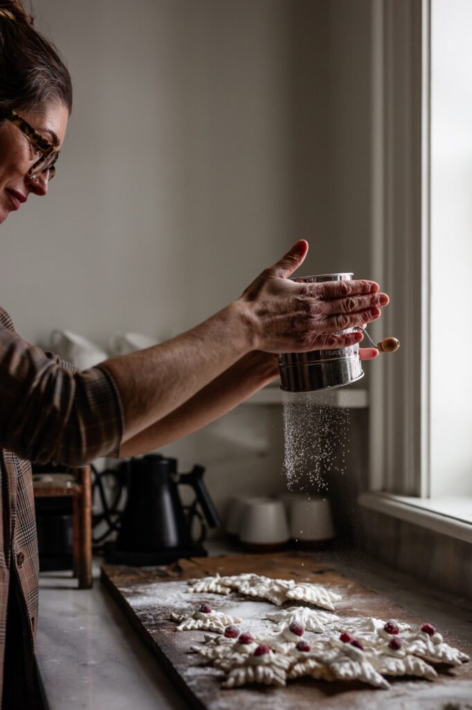 A woman dusts powdered sugar over a table full of white holiday cookies.
