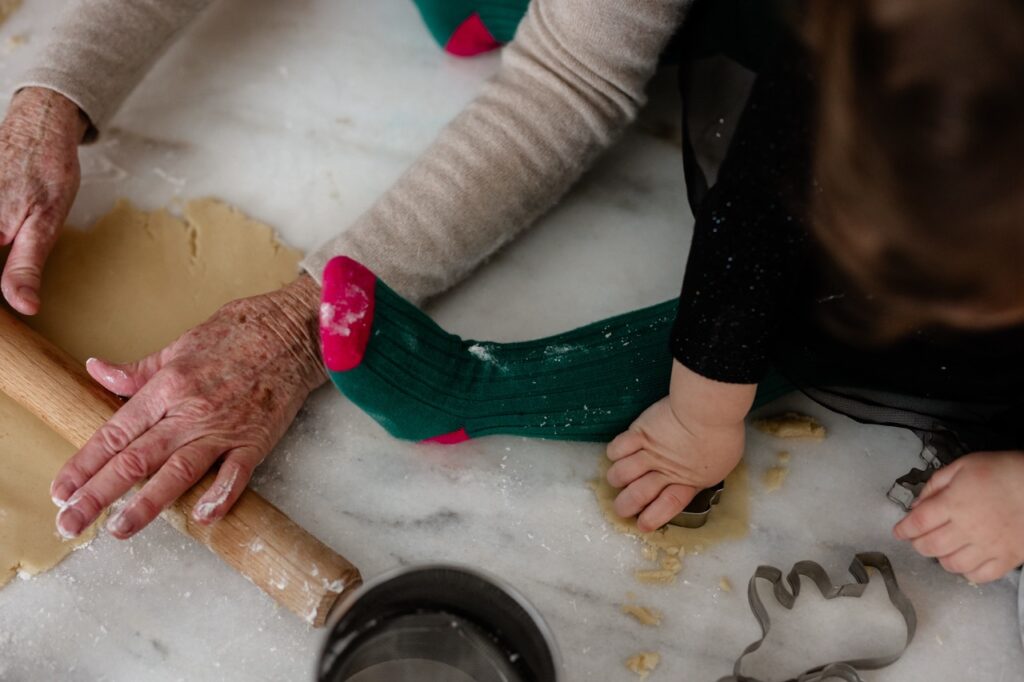 A woman rolls out cookie dough as a young girl in green socks sits beside her.
