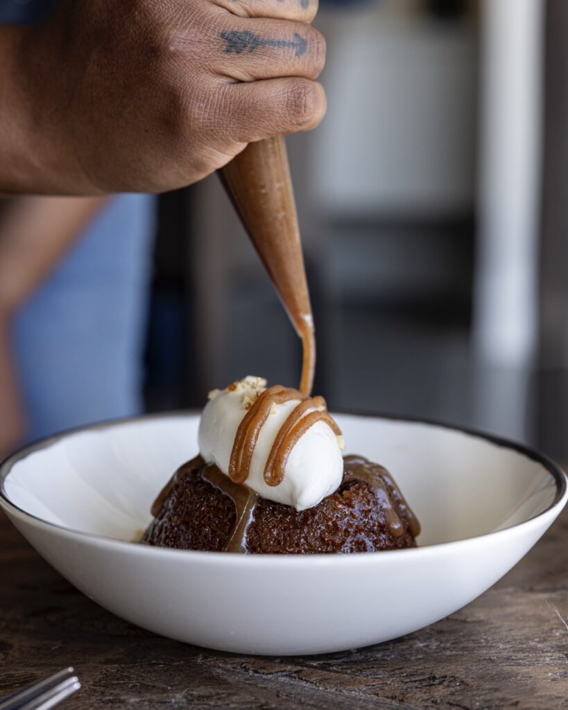 A woman pipes a sauce onto a servings of Malva Pudding in a white bowl.