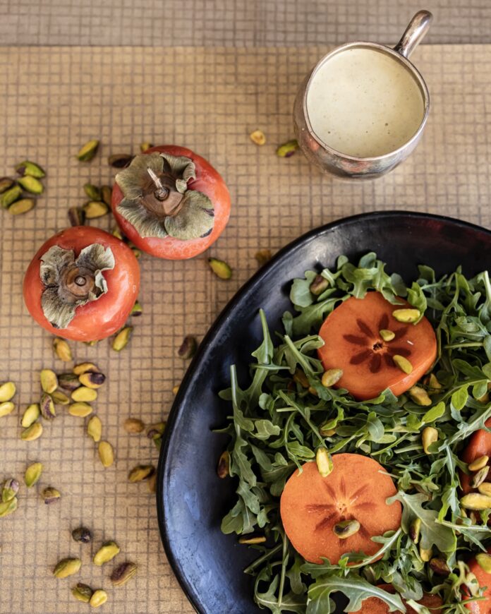 Using Persimmons in the Winter A winter salad of arugula and persimmons sits on a blue plate with whole persimmons, seeds, and a dressing container nearby.