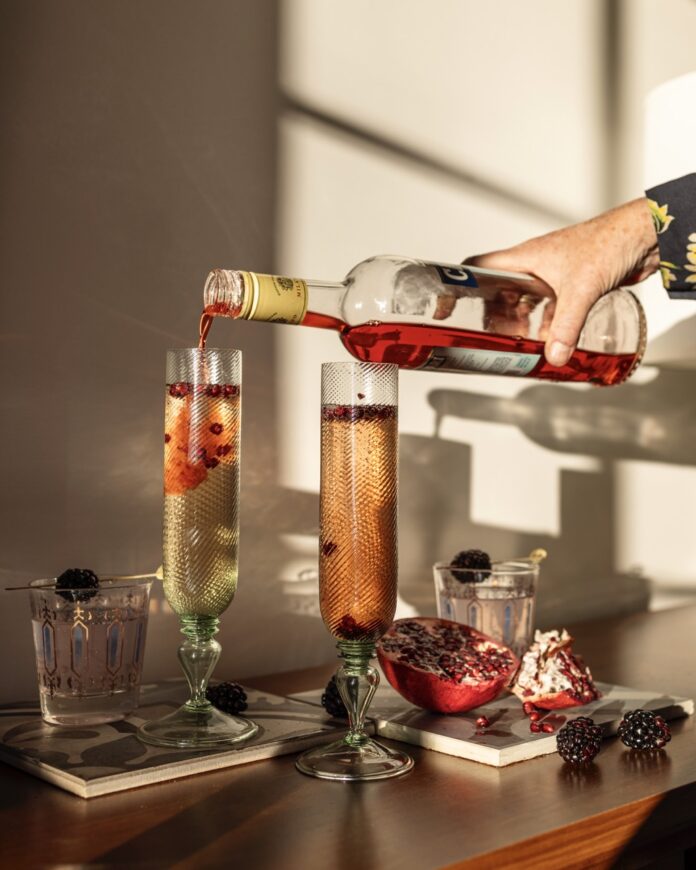 A person pours prosecco on top of a pomegranate cocktail in two tall glasses.