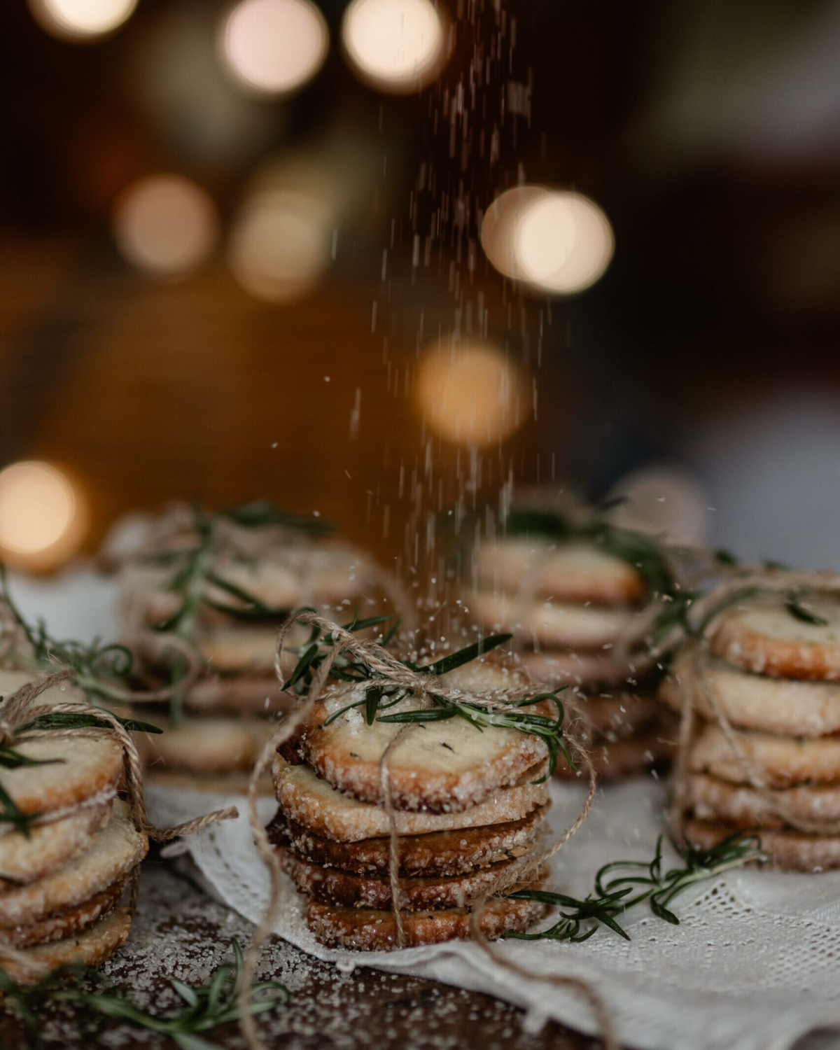 Stacks of Rosemary Butter Cookies sit wrapped in twin while someone sprinkles sugar overtop.