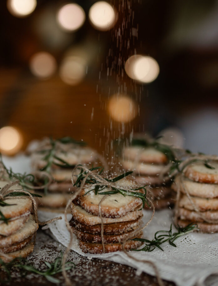 Rosemary Butter Cookies Stacks of Rosemary Butter Cookies sit wrapped in twin while someone sprinkles sugar overtop.