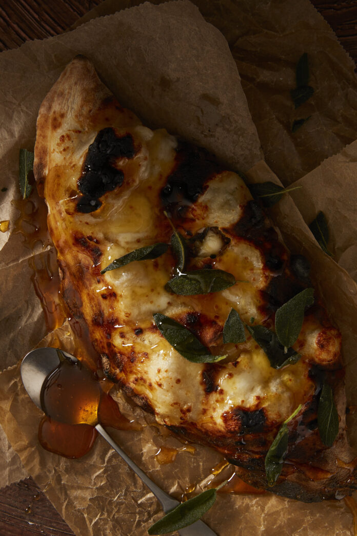An oblong calzone sits on a table stuffed with sausage, squash, and topped with sage leaves and honey.