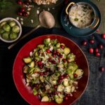 A red bowl filled with shaved Brussels sprouts topped with cranberries, almonds and shredded cheese. Small accompanying bowls contain additional Brussels sprouts, shredded cheese and almonds.