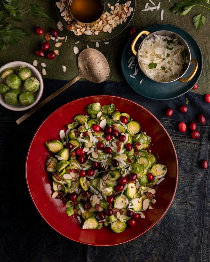 A red bowl filled with shaved Brussels sprouts topped with cranberries, almonds and shredded cheese. Small accompanying bowls contain additional Brussels sprouts, shredded cheese and almonds.