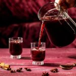 A pitcher filled with a dark red sangria cocktail garnished with cinnamon sticks and star anise pods, as described in the recipe for the "Spice is Nice Sangria" batch cocktail.